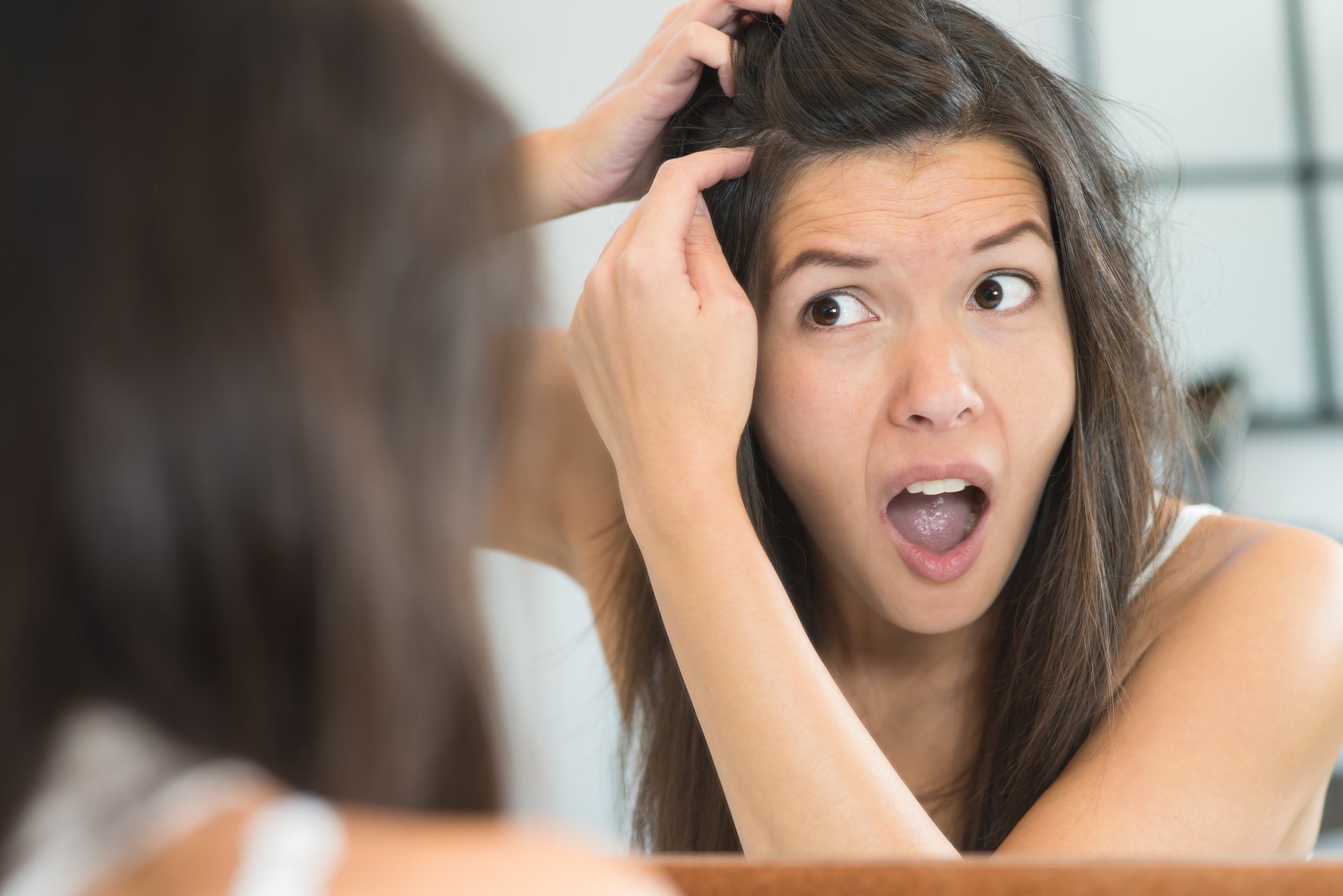 Horrified young woman looking in the bathroom mirror staring open mouthed at the first grey hair on her scalp, a first sign of ageing, or noticing that she is suffering from dandruff